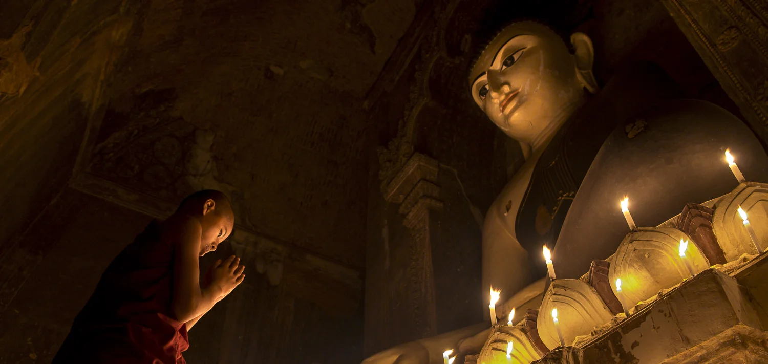 Large candle lit buddha gazing down at novice monk in a temple in Myanmar